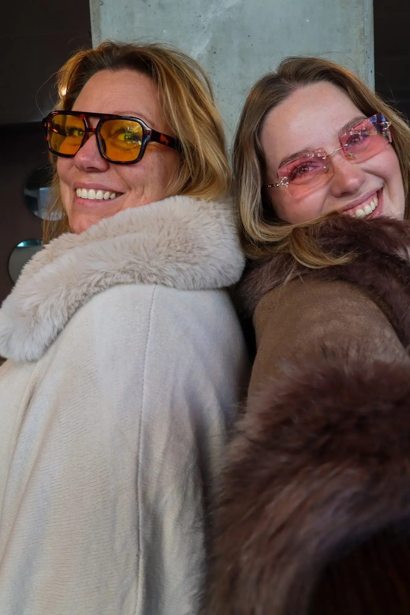 Laila og Amalie i pelsjakker og solbriller, smilende ryg mod ryg